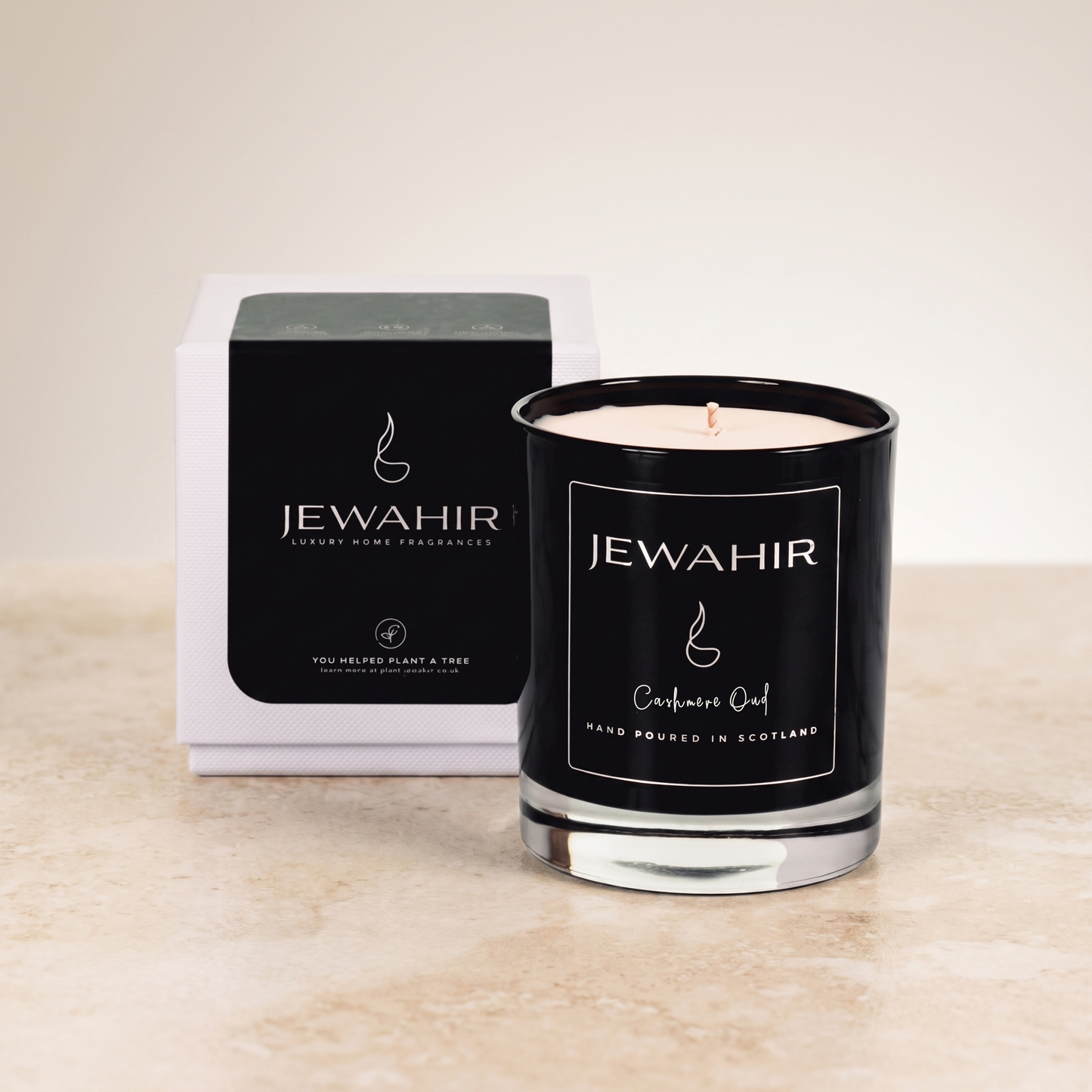 A black glass jar candle with a white wick and cream-colored wax sits in front of its white and black retail box on a light beige stone surface against a neutral beige background. The label reads 'JEWAHIR' and 'Cashmere Oud'.