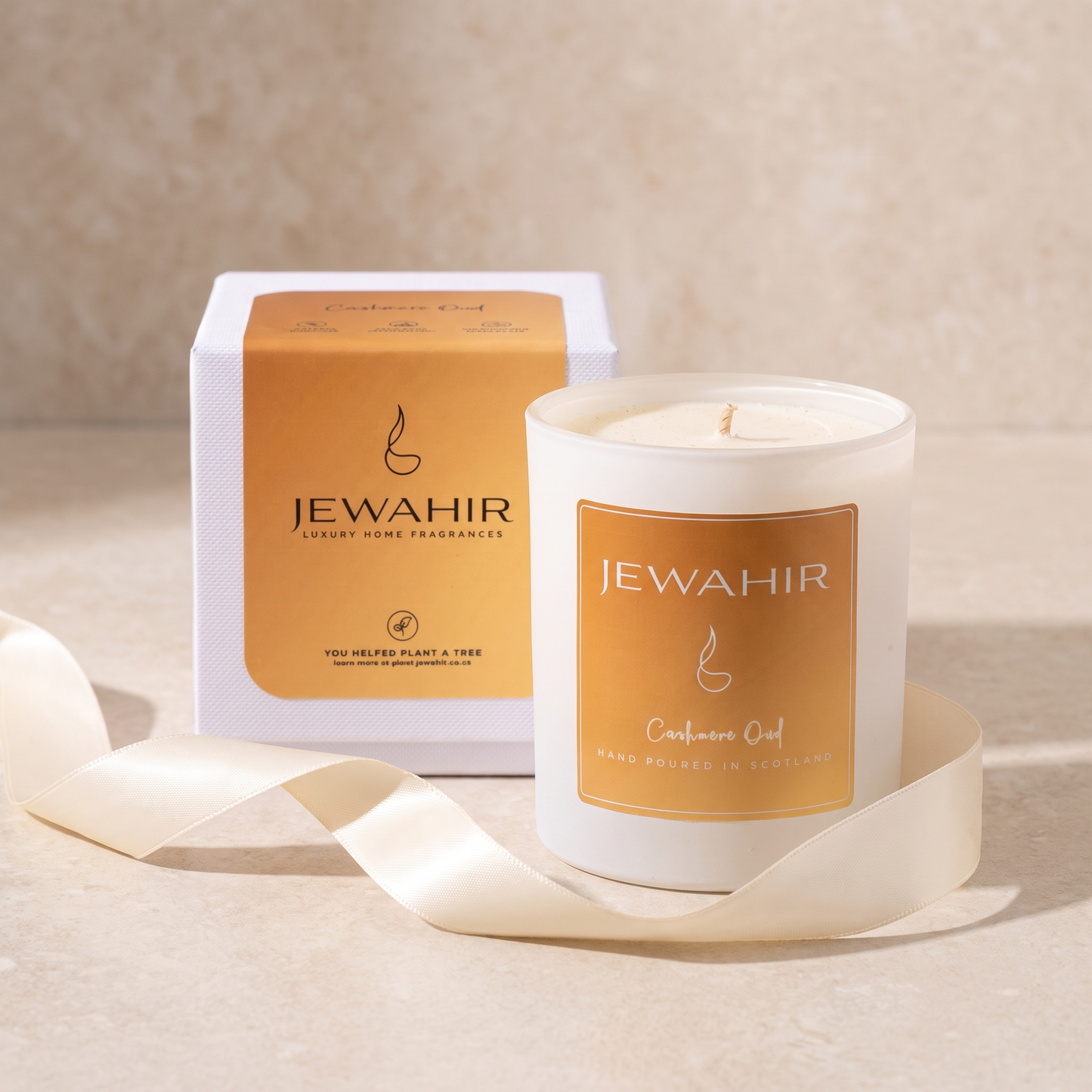 A white glass jar candle labeled JEWAHIR Cashmere Oud sits in front of its white and gold box, with a cream ribbon draped around the base on a warm beige surface.